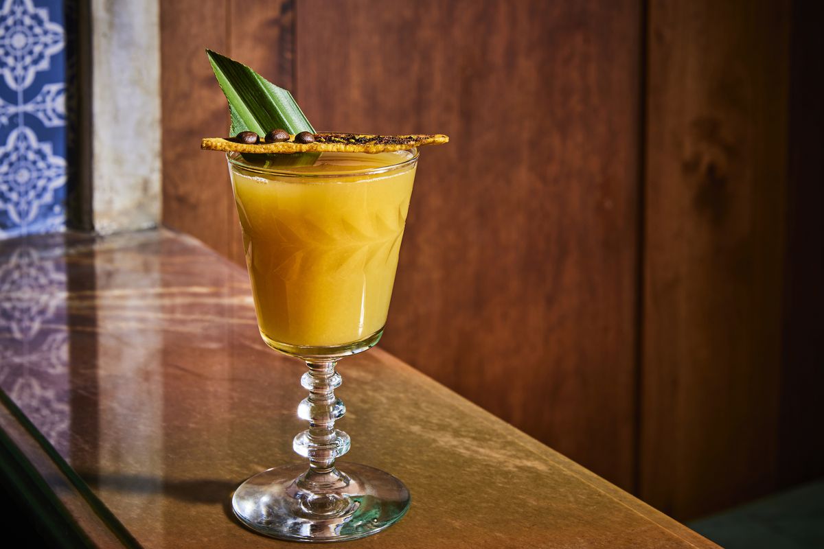 A yellow cocktail with floral flourish and greenery poking out on top, set on a wooden table at new LA restaurant Que Barbaro.
