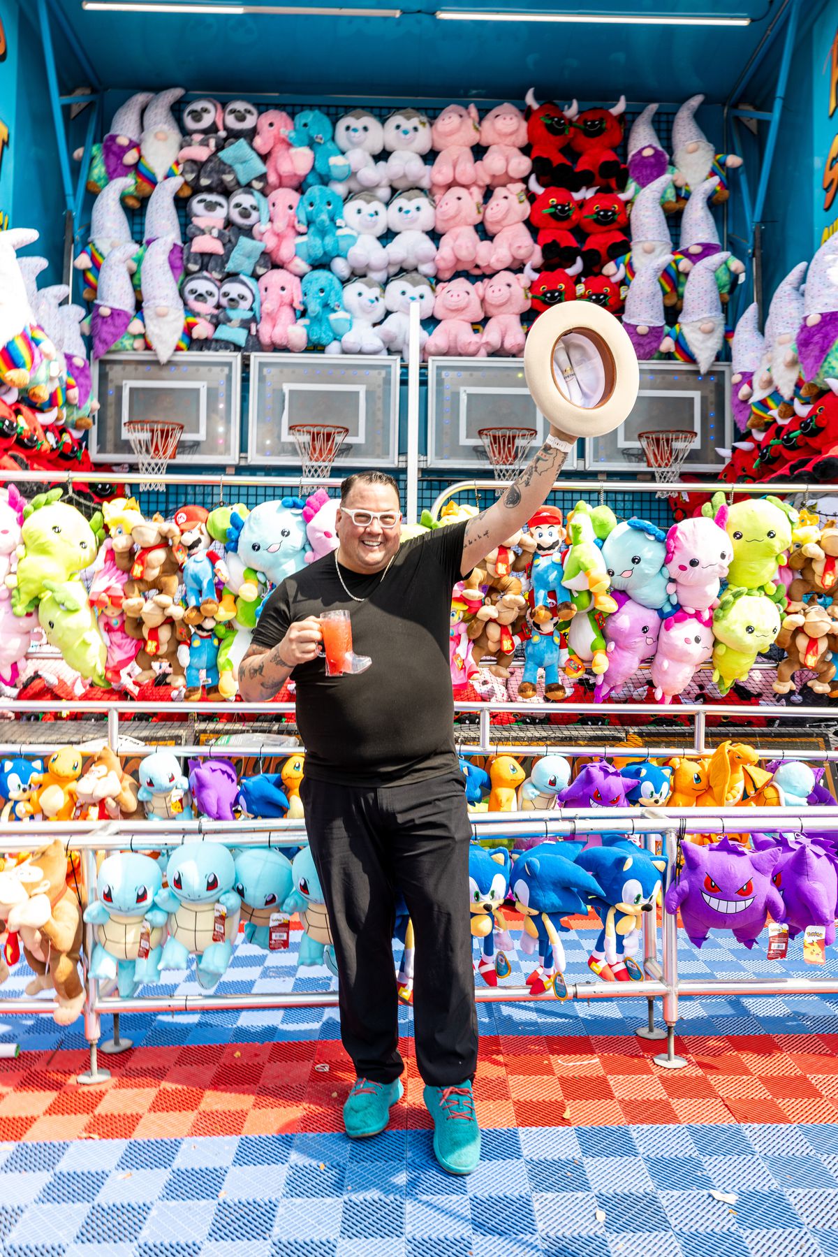 Celebrity chef Graham Elliot stands in front of a fair game with loads of colorful stuffed animals. He’s dressed all in black and waves a cowboy hat with his right hand. In his left, he holds a red drink in a clear plastic boot.