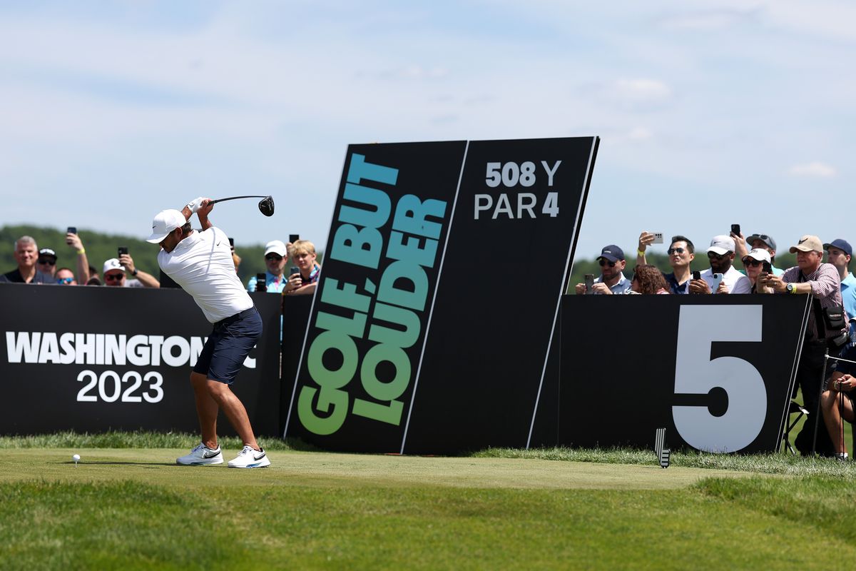 A golfer taking a swing in front of a sign that reads “Golf, but louder” and a crowd behind a barricade.