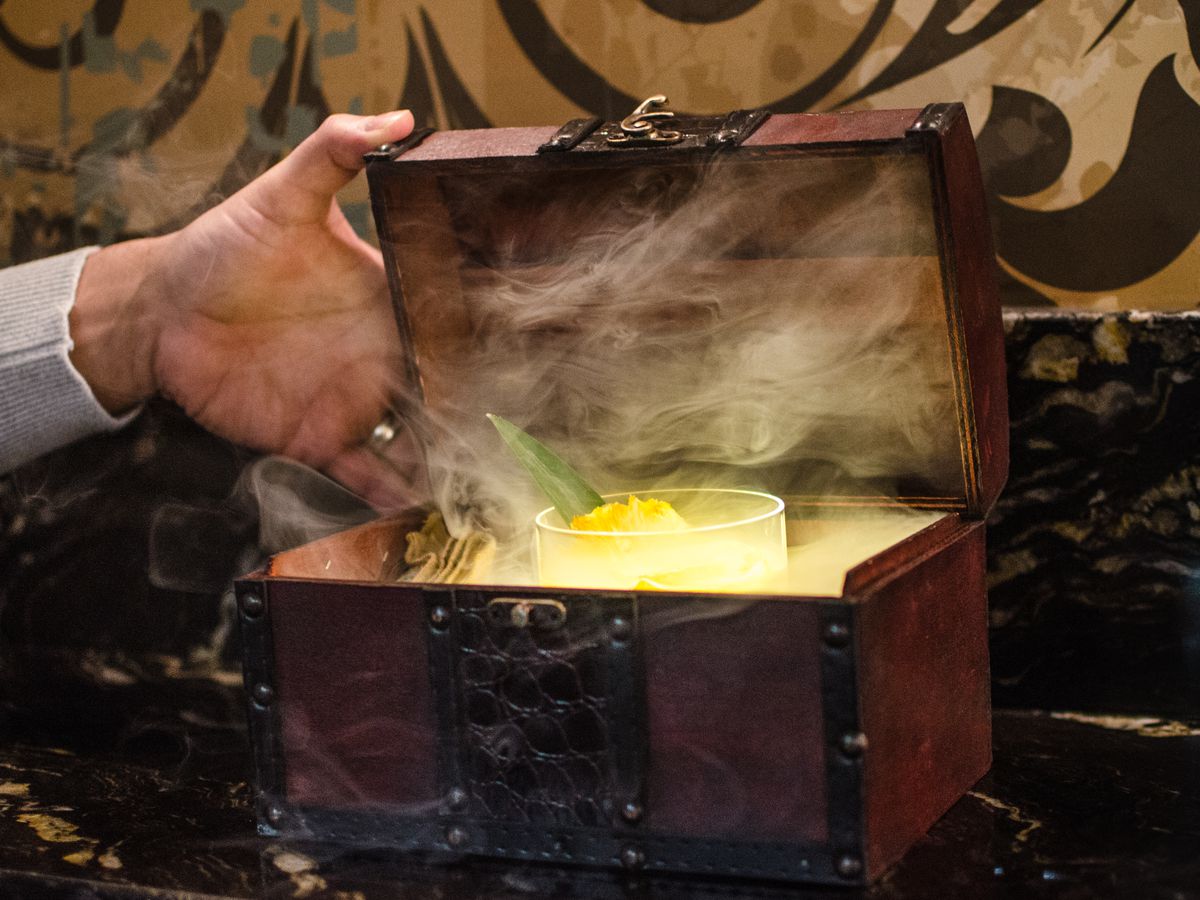 A hand opens a small treasure chest to reveal a yellow cocktail in a cloud of smoke.