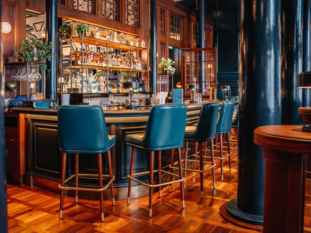 A luxe bar with blue leather stools, back-lit shelves of spirits, and rich wood. 