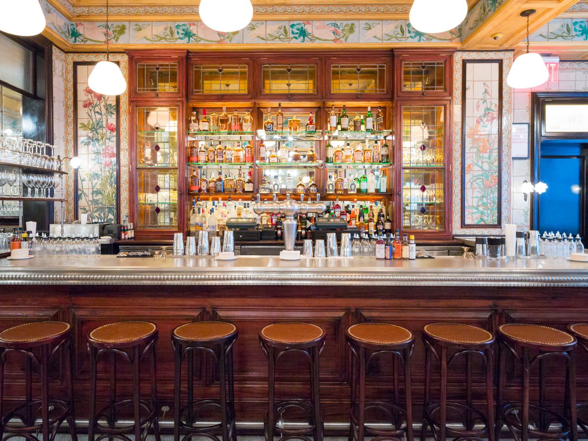 Brown barstools stand against a long wooden bar, where four rows of alcohol bottles are backlit