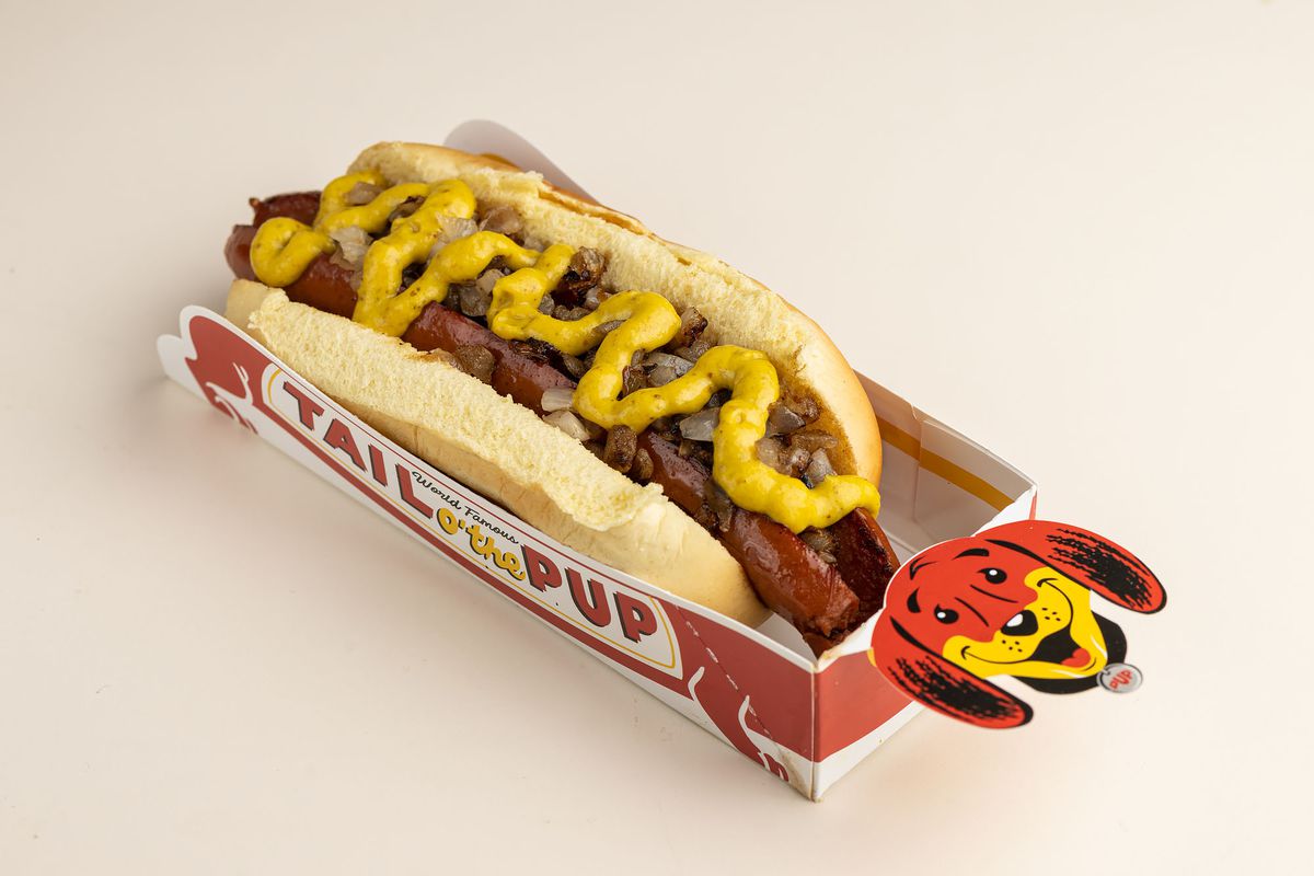 A hot dog with mustard swizzled on top inside of a red cardboard sleeve.