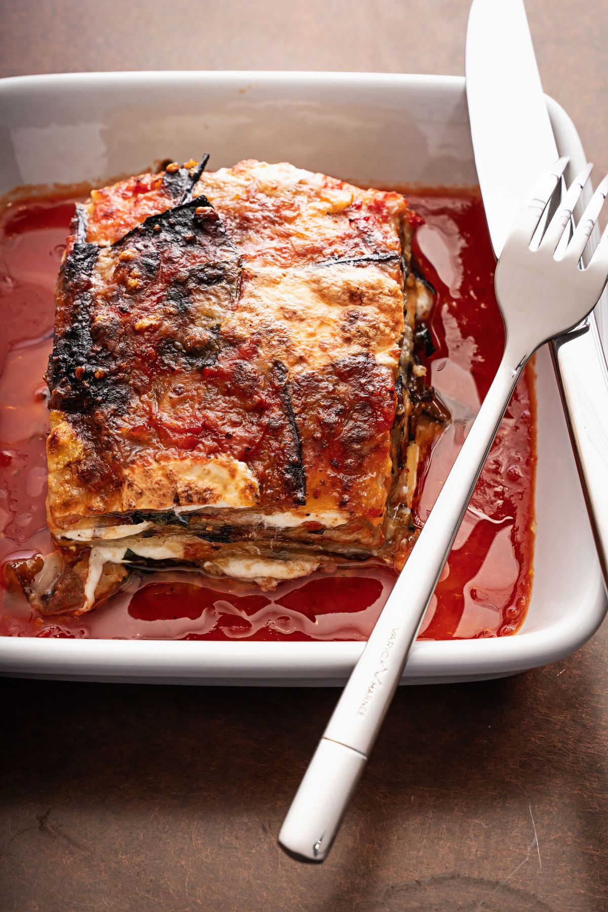 A layered eggplant Parmesan in a white ceramic tray with a knife and fork to the right.