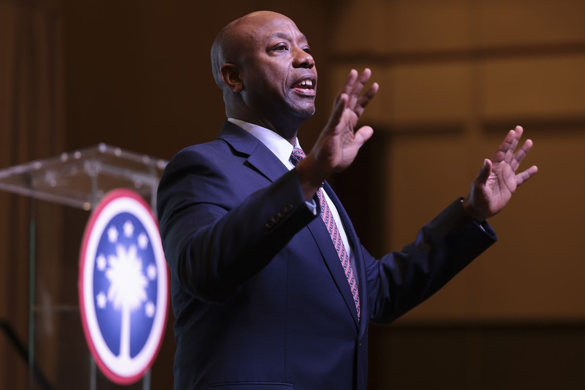 Tim Scott is pictured in mid-speech, with both palms raised in a gesture. He wears a navy suit and red tie.