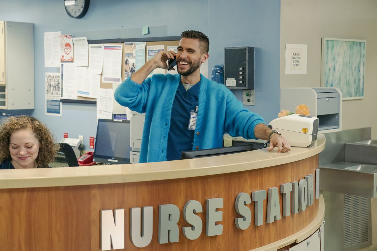 Josh Segarra as Lance Arroyo in The Other Two, standing at the nurse station in a hospital in scrubs and a teal cardigan, smiling while he talks on the phone, handsomely.