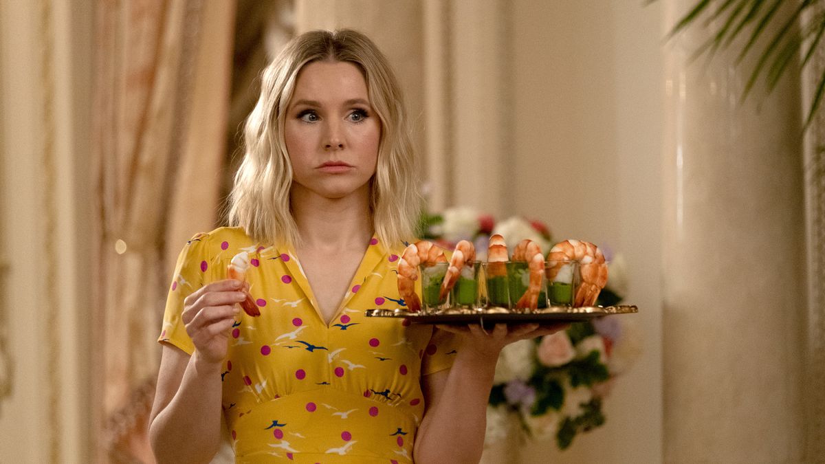 Kristen Bell, wearing a bright yellow patterned dress, holds up a tray of shrimp cocktails while looking dubious and dissatisfied.