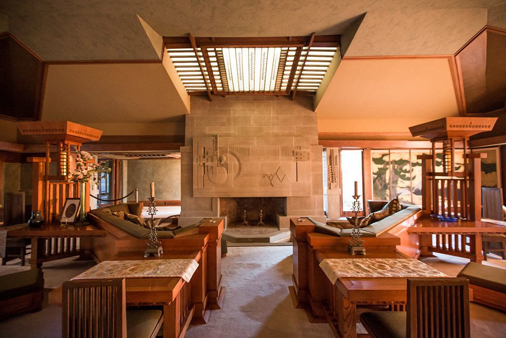 The Hollyhock House by Frank Lloyd Wright. The interior has couches, tables, a fireplace, and a skylight with wooden slats. The wall above the fireplace is brown brick. There is a tan floor.