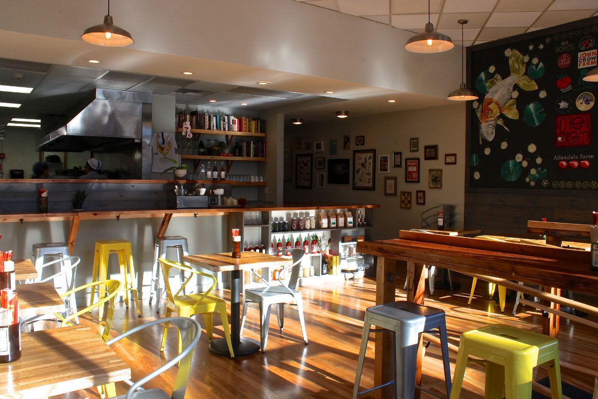 A casual restaurant interior with sunlight streaming in. There are light wooden surfaces, an open kitchen, and yellow and white metal chairs.