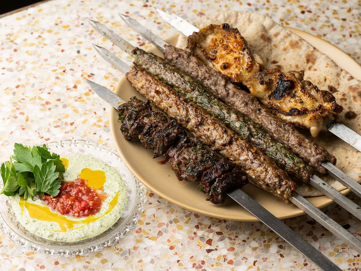 A wide shot of five metal skewers of grilled meat, plus bread, on a plate at a restaurant.