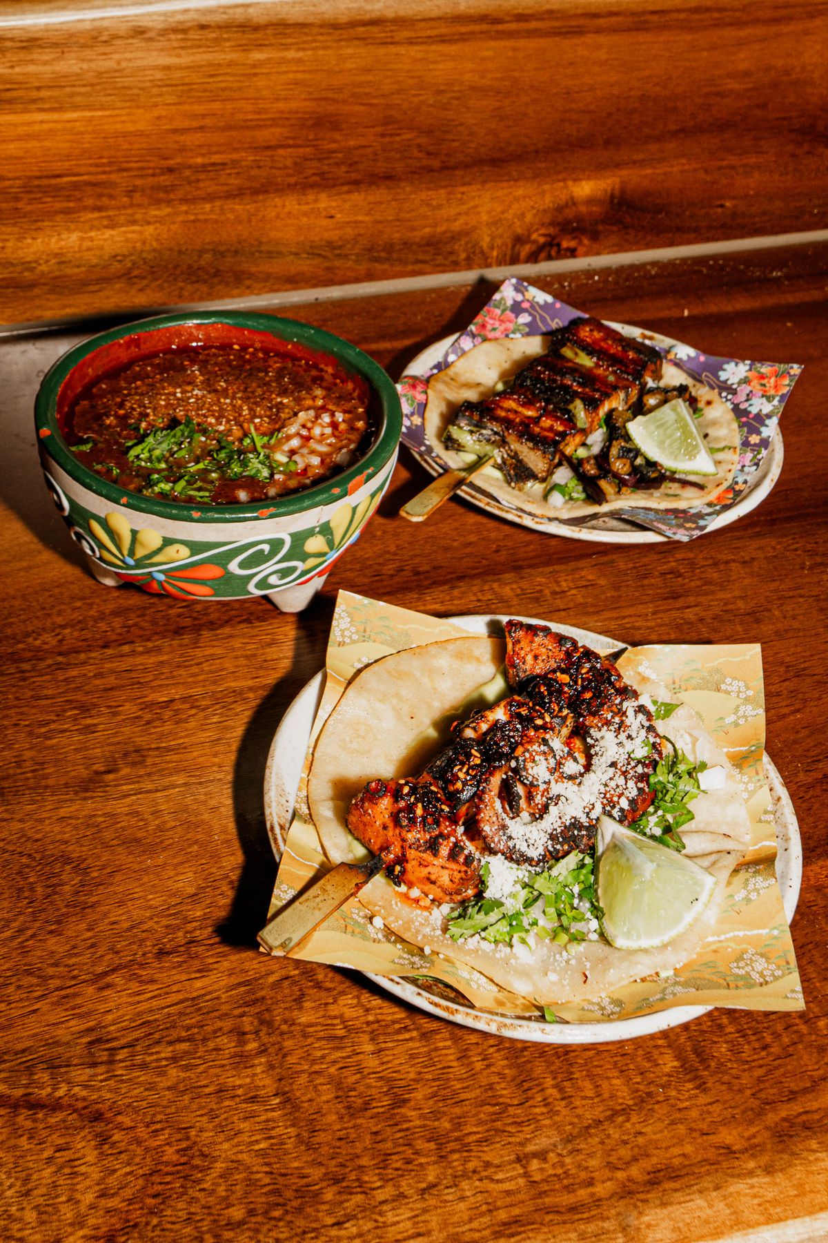 A bowl of broth next to two plates of tacos with skewered meats.