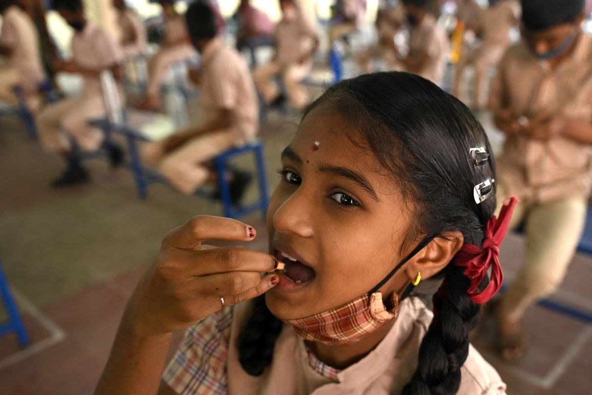 A young girl with two braids and a plaid mask below her chin puts a small pill into her mouth, surrounded by a group of classmates.