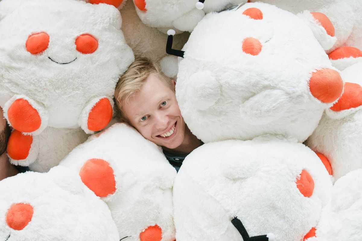 A caucasian man with blonde hair smiles out from a pile of plush toys shaped like the Reddit logo.