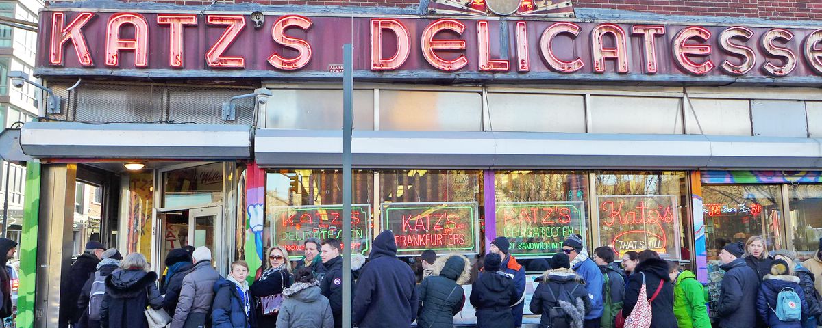 A line of people outside the deli, the front window of which is filled with neon signs