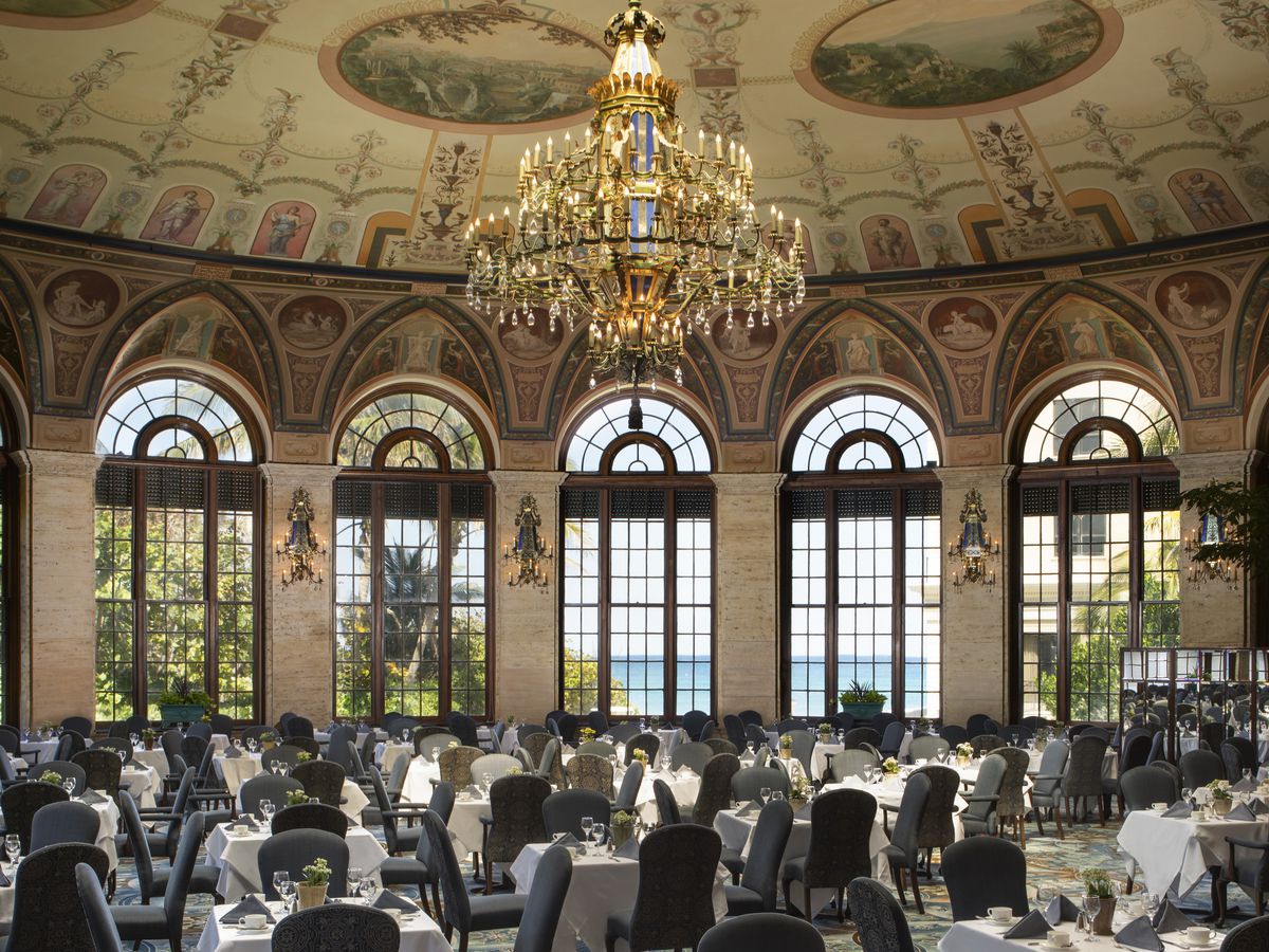 This is a picture of a round dining room at The Breakers in Palm Beach. There are tall, arched windows overlooking the Atlantic ocean. There are dining tables under a large crystal chandelier.