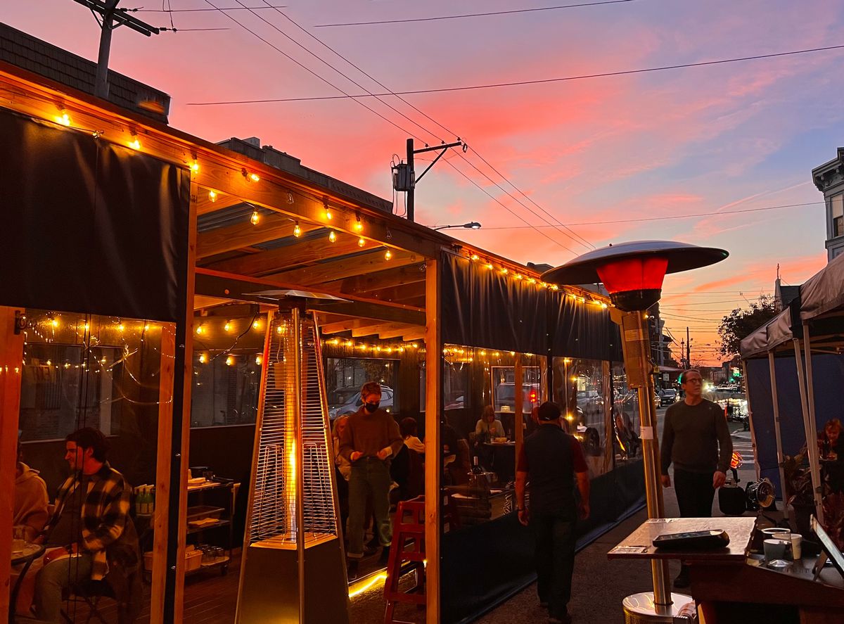 A streetery setup with heated patio lamps and diners at tables inside booths with a pink sky above them.