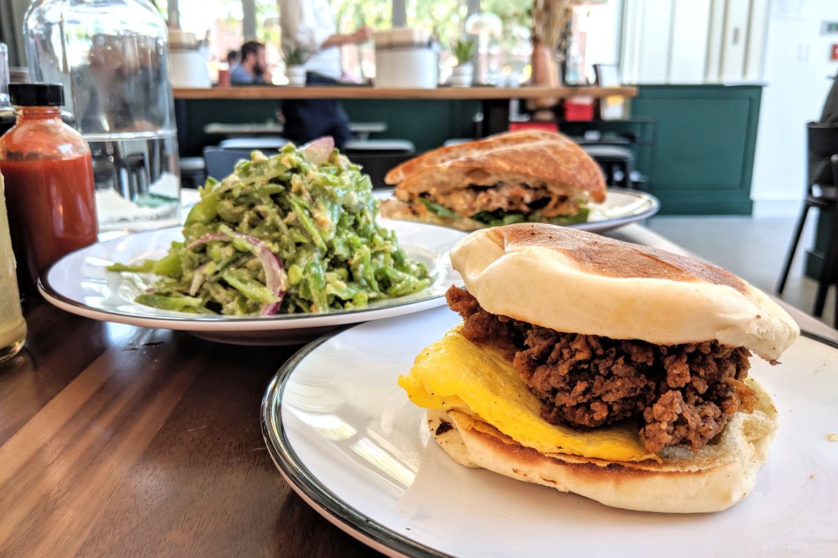 A fried chicken sandwich served on a Portuguese bun is in the foreground of the photo, with a stacked-high sugar snap pea salad and a roasted cauliflower sandwich in the background. Shot at Shy Bird in Cambridge.