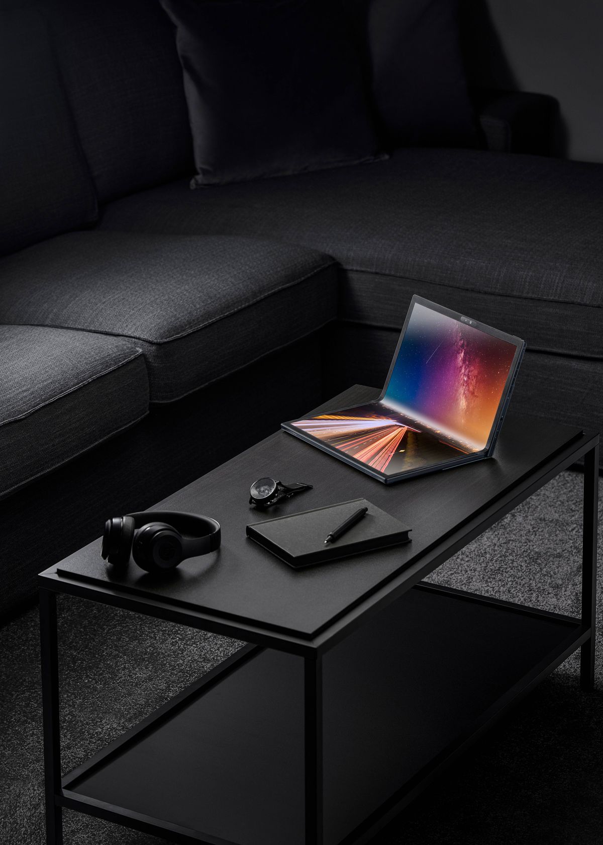 The Asus ZenBook 17 Fold OLED is in clamshell mode on a black coffee table with a black couch behind it.  Next to it is a black watch, a black pair of headphones, and a closed black notebook with a black pen on top.  The screen displays a multicolored background.