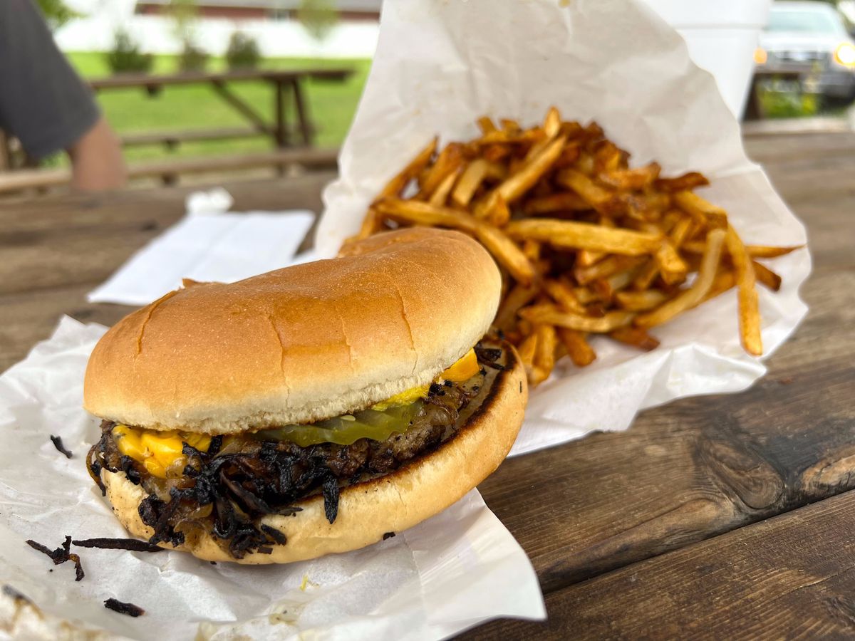 A burger with deeply singed onions on a picnic table beside a pile of fries.