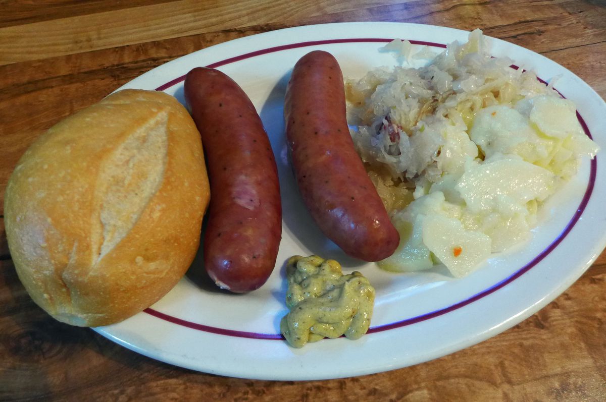 Two sausages on a plate with potato salad and a roll.