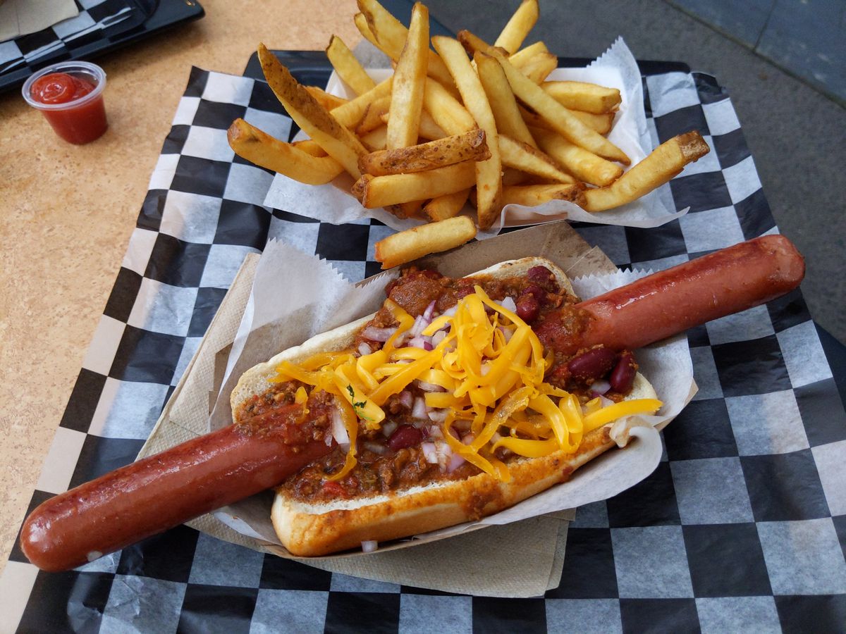 A long hot dog in a short bun on a checked paper with french fries in the background.