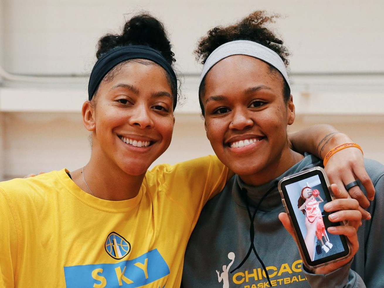 Two Black women standing next to each other, smiling, while the woman on the right holds her phone up to the camera so a doll is visible on the screen.