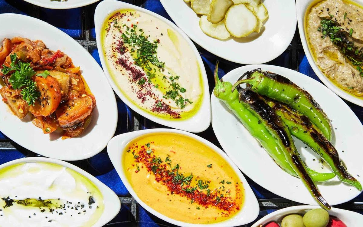 From above, a table full of colorful dishes, including whole grilled peppers, dips, and pickles.