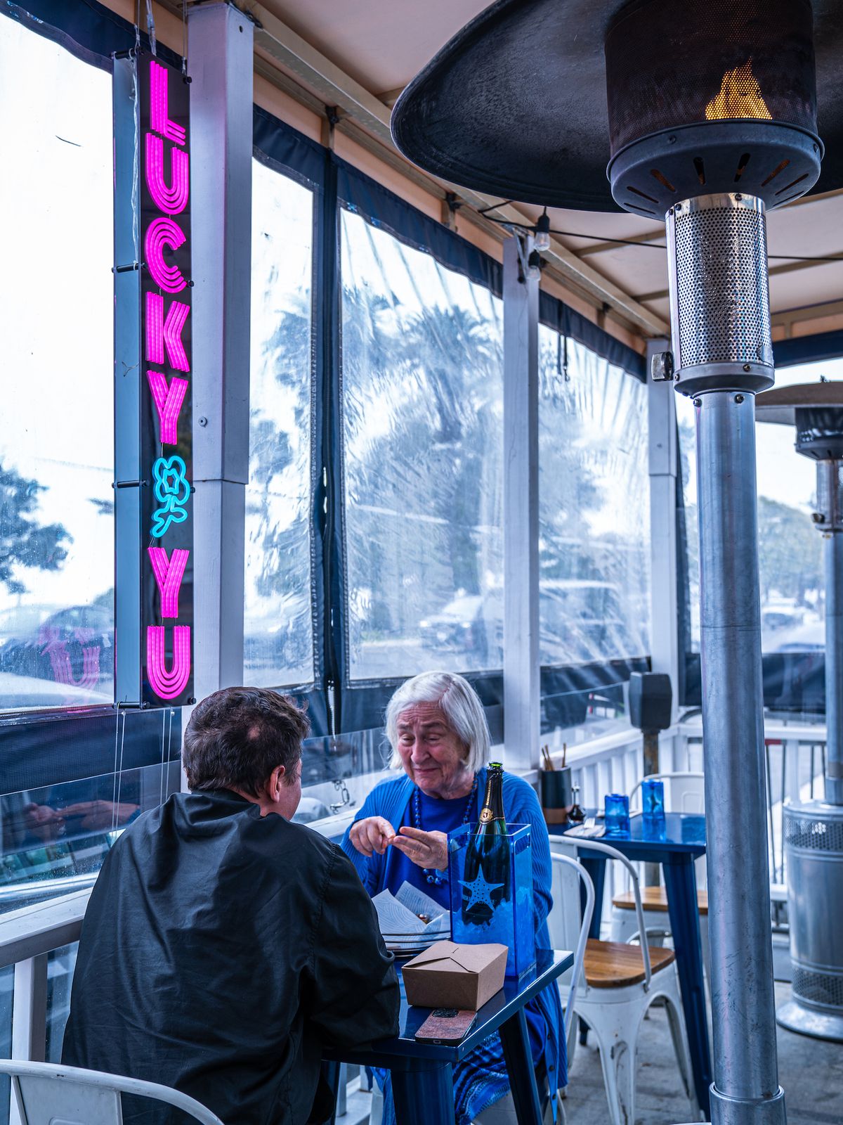 Two customers sit beneath a neon sign and next to a rain protector at a stormy daytime seafood restaurant.