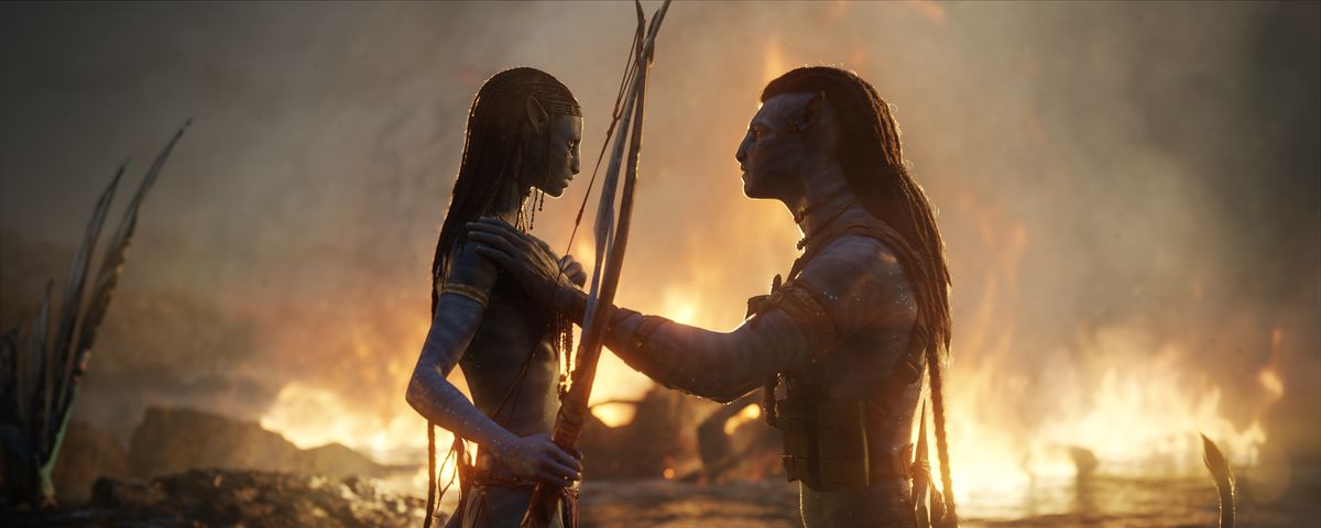 Na’vi warriors Neytiri (Zoe Saldaña) and Jake Sully (Sam Worthington) face each other in front of a landscape on fire in Avatar: The Way of Water