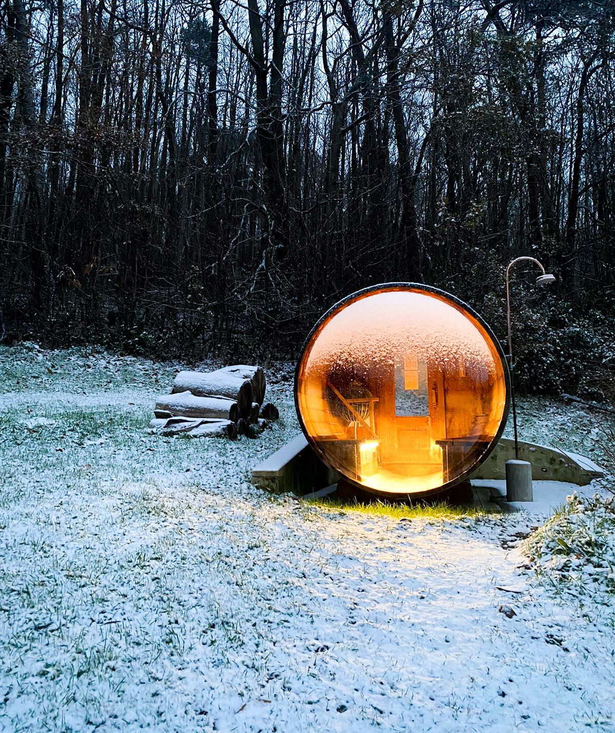 A single-room sauna with a large glass wall lit up in the middle of a snowy forest scene. 
