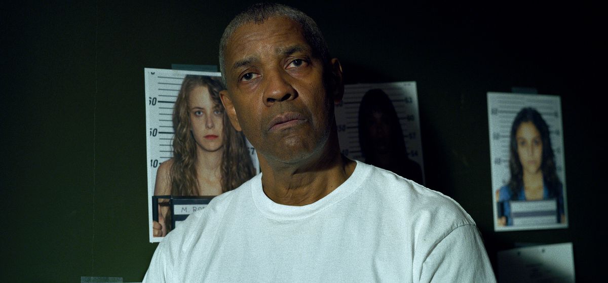 Denzel Washington wears a white t-shirt and stands in front of a green wall with pictures of murdered women in The Little Things