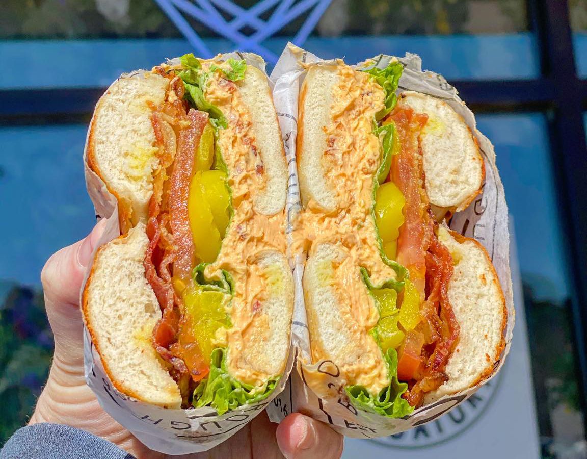 A bagel sandwich cut in half to reveal meat, vegetables, and flavored cream cheese, held in front of a neon ‘open’ sign