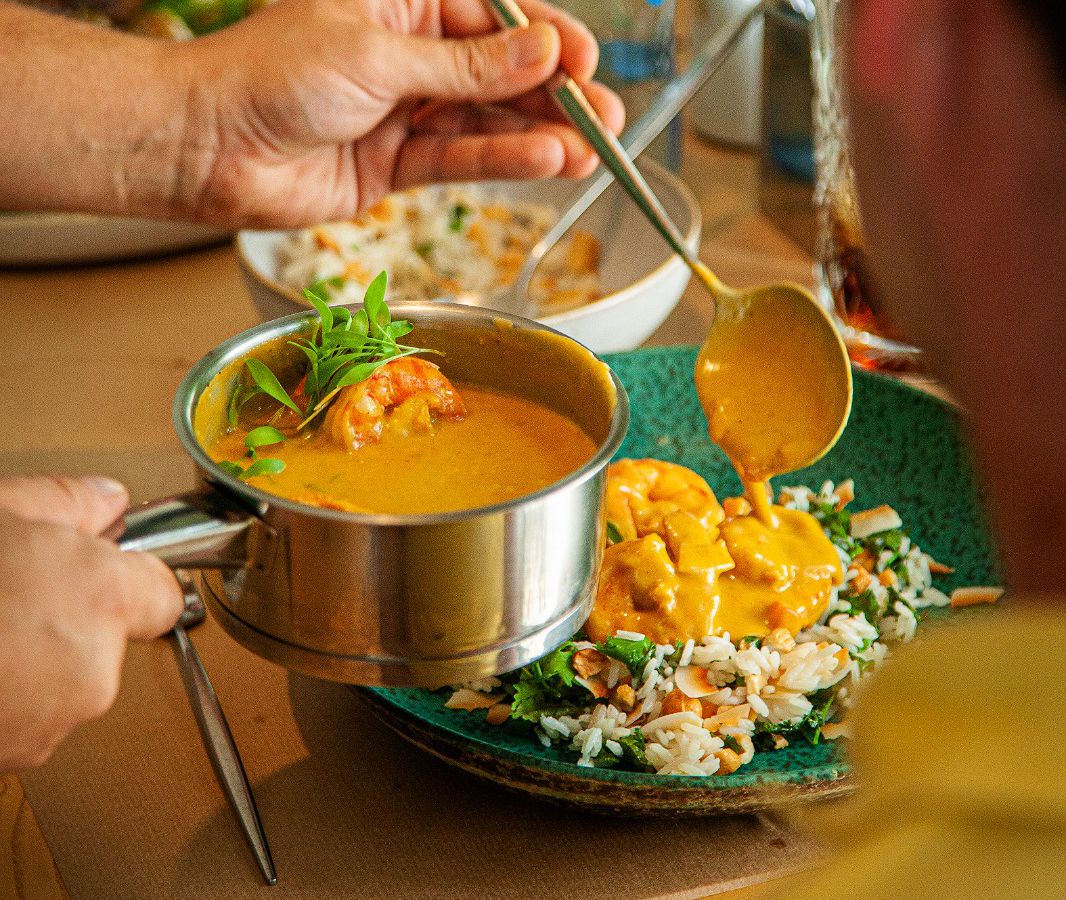 A server ladles sauce from a saucepan of curry, where a shrimp bobs, onto a plate of rice and vegetables.