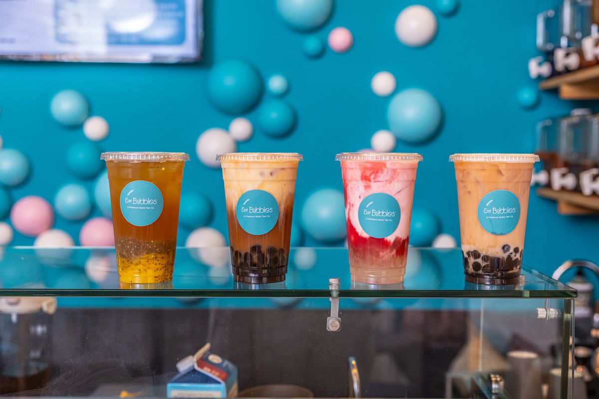 Four different flavors of boba lined up horizontally on a glass counter set against a bright, turquoise colored wall