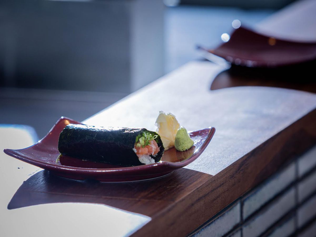 A curving plate sits on a bar with a seaweed-covered roll of temaki placed on top with ginger and wasabi on the side.