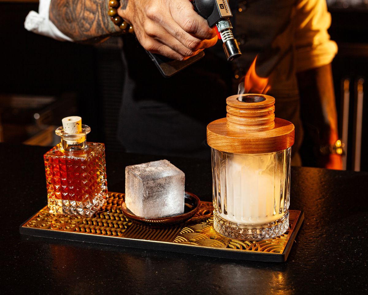 A bartender uses flame to create smoke on top of a cocktail.