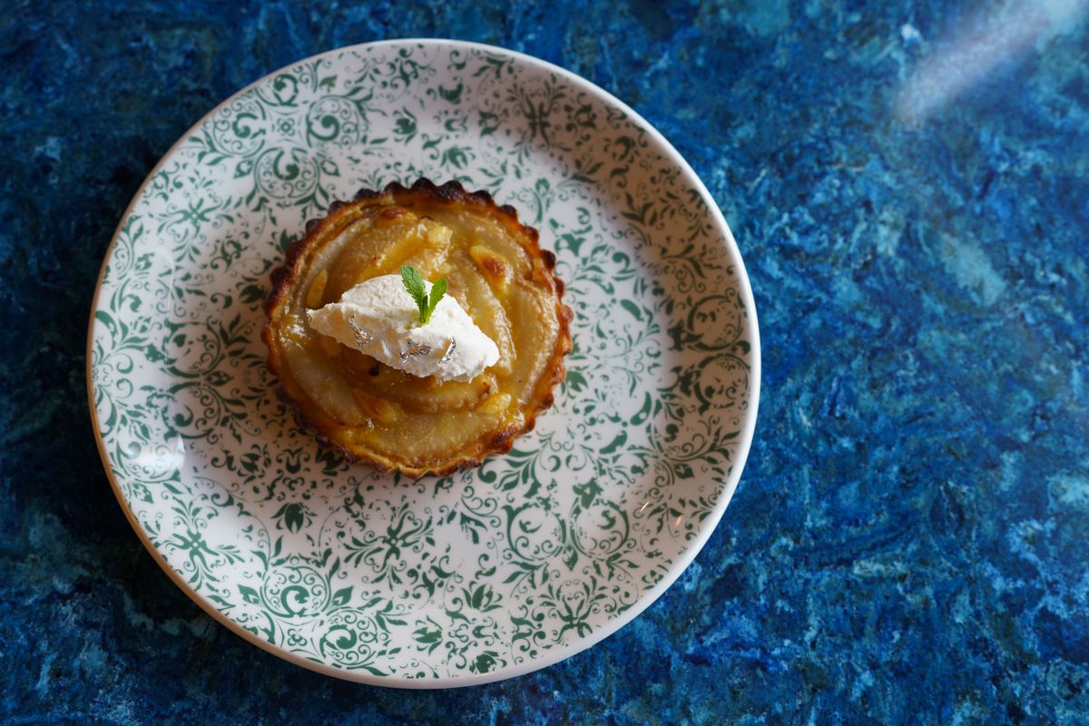 A pear tart topped with whipped cream and a sprig of mint on a white and green patterned plate resting on a blue table.