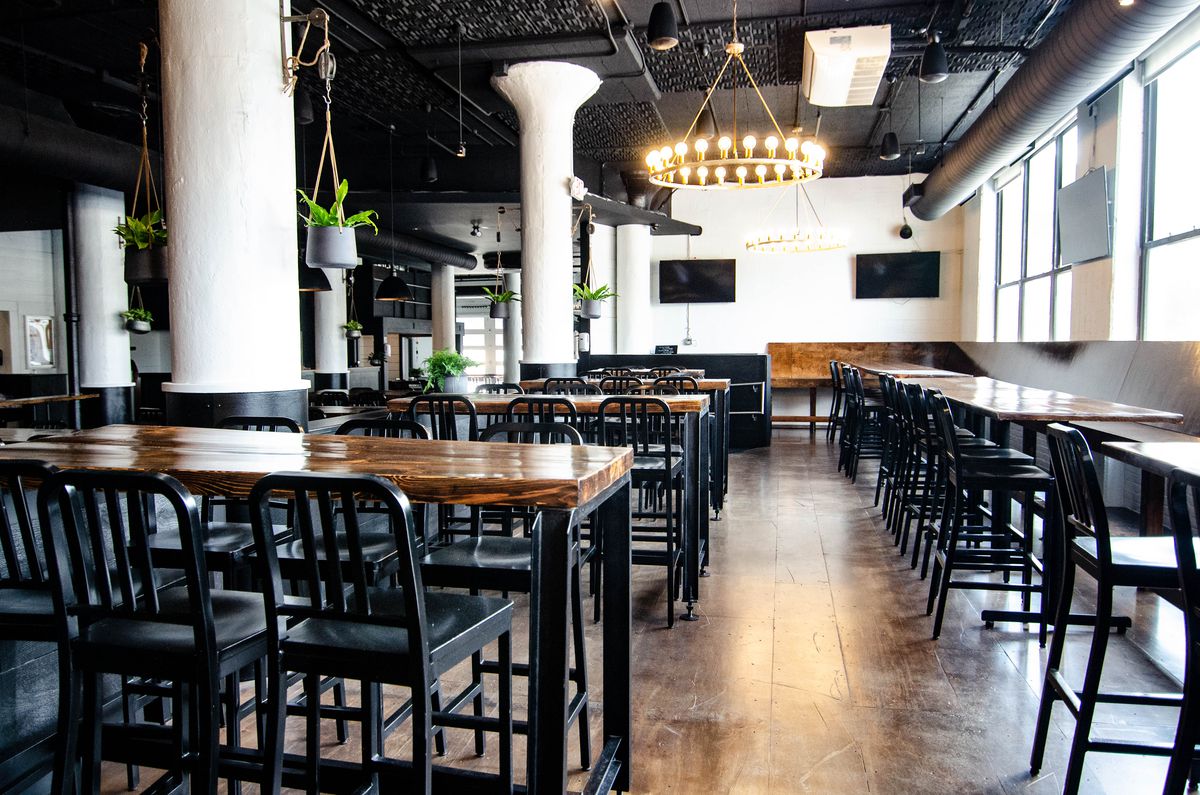 Restaurant interior features narrow high-top tables with a glossy wood finish, black chairs, and hanging plants in a largely black-and-white space.