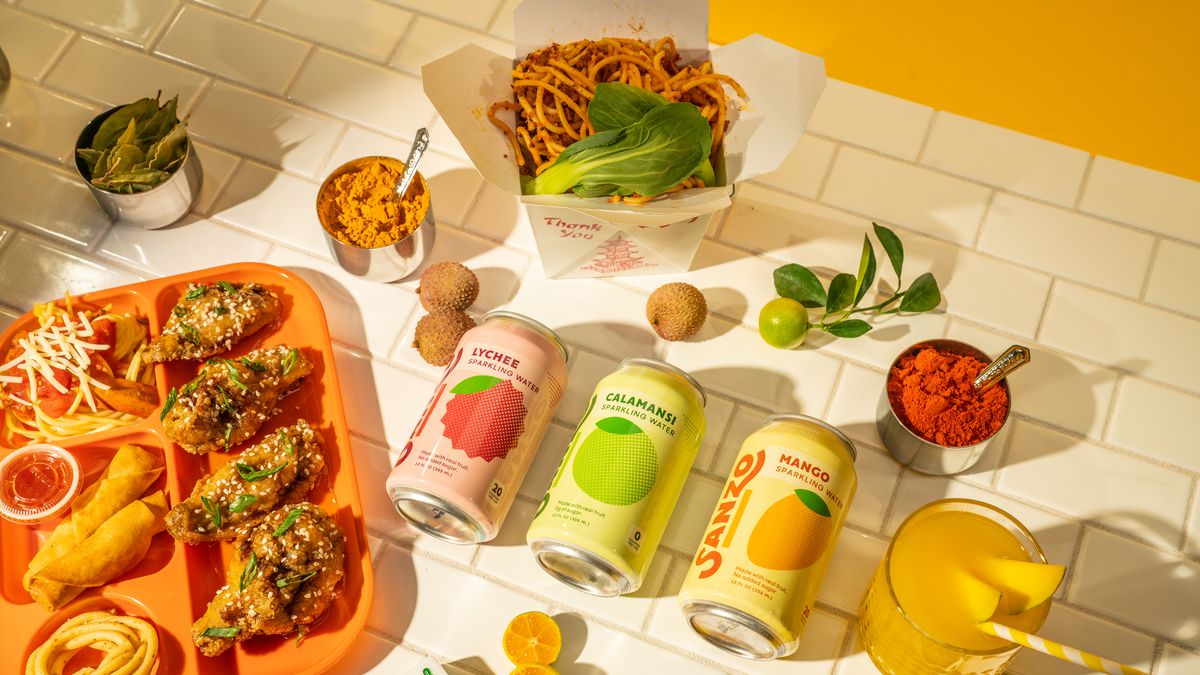 Three cans of Sanzo sparkling water sit on a table with items like fresh fruits, fried chicken, noodles, and spices. 