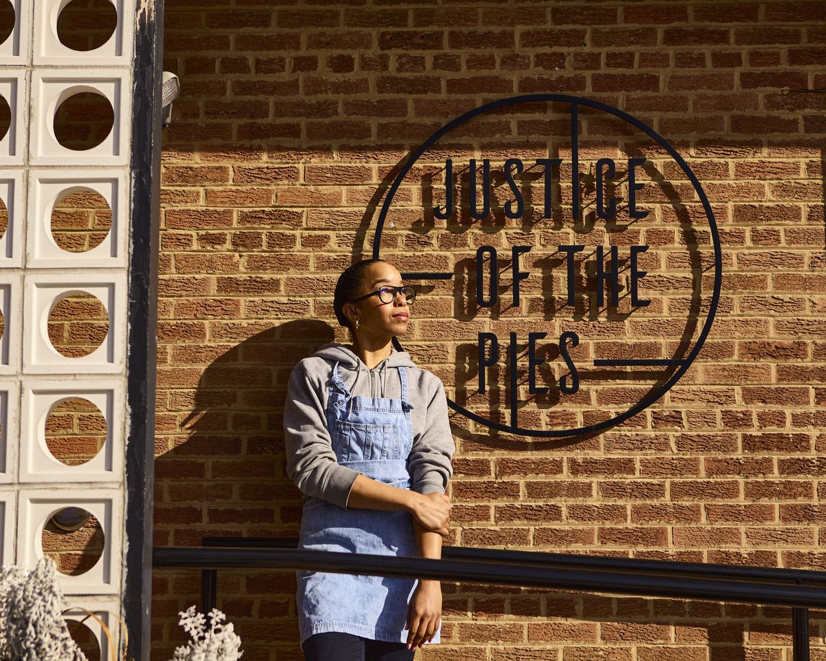 Chef Maya-Camille Broussard dressed in a blue apron and sweatshirt in front of her shop.