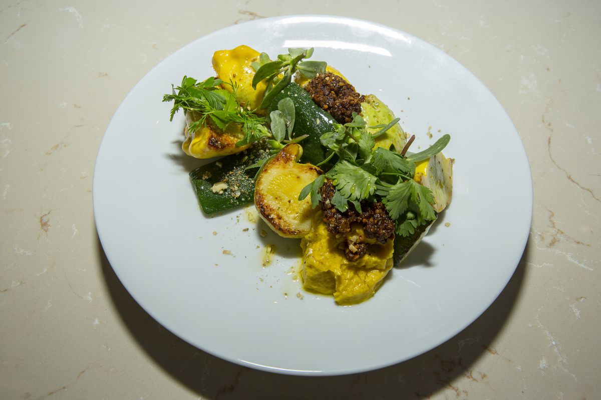 A white plate contains charred green summer squash with smooth yellow squash hummus, brown salsa roja, and green parsley. 