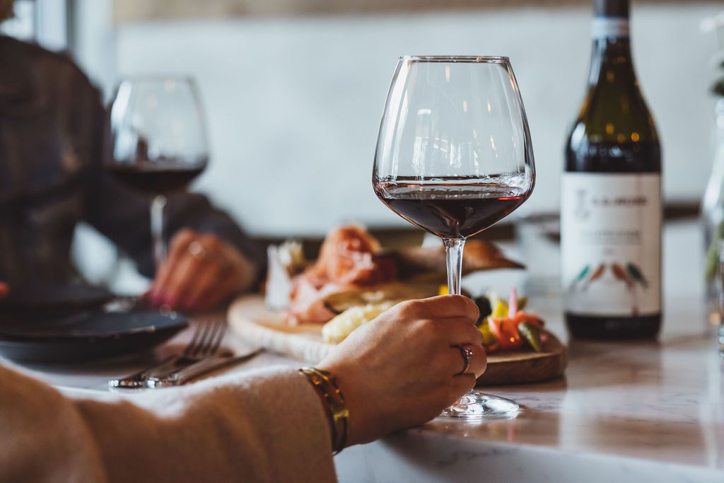 A hand holds a glass of red wine in front of a platter of charcuterie, with a bottle of red wine in the distance. The two guests, out of frame, are sitting at the bar at Normandie in Southeast Portland.