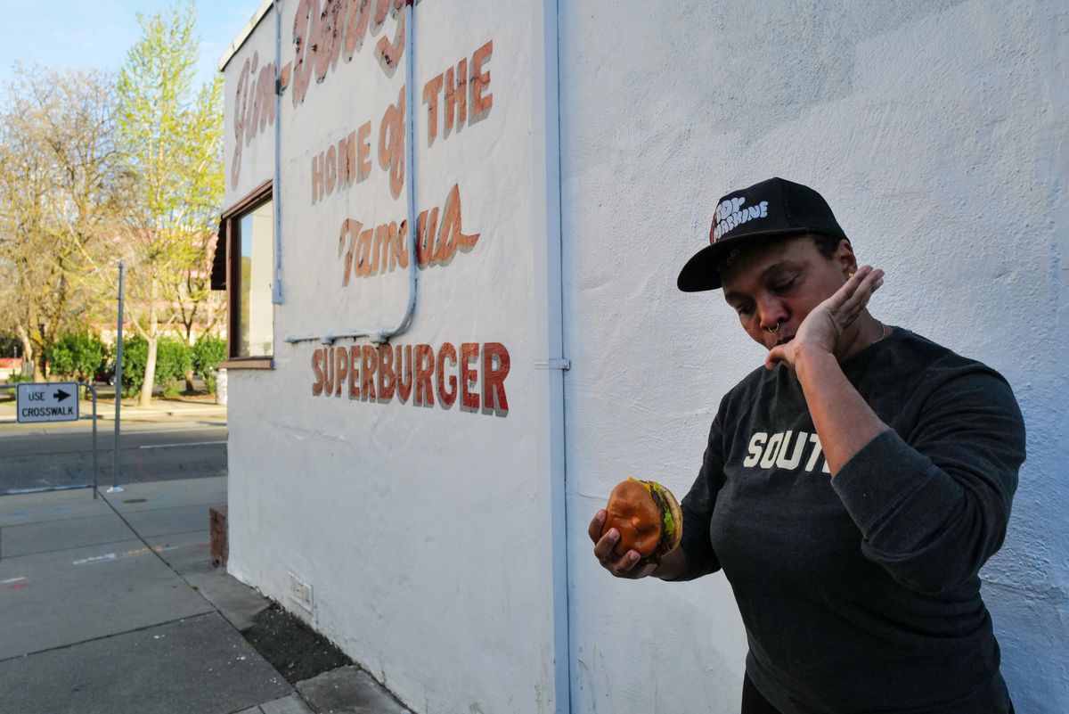 A woman wearing a t-shirt that says “South” eats a burger outside a white building.