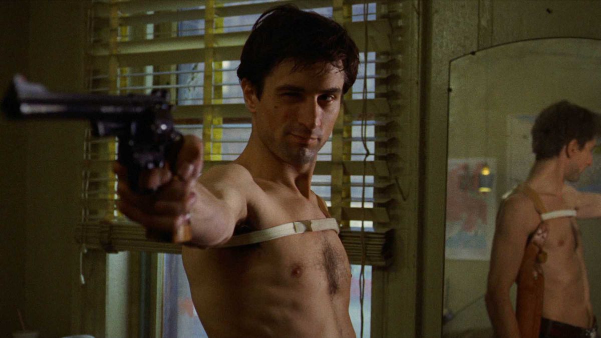 A white man stands, bare-chested, with a holster, pointing a handgun.