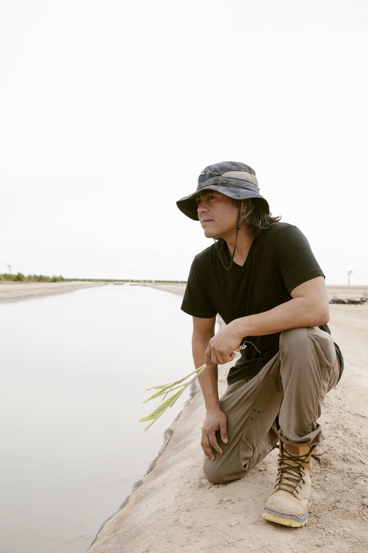 A man in a slouchy hat holds a stalk of greenery while kneeling beside a channel of water.