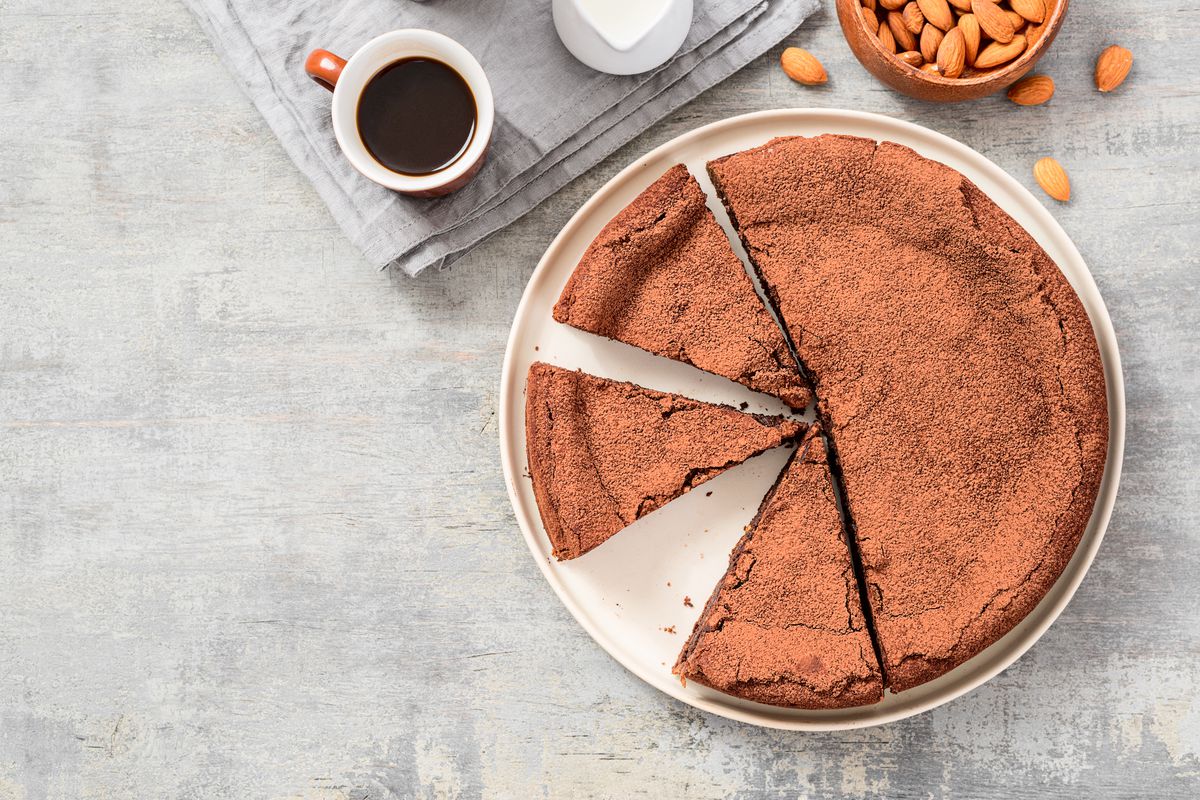 A flourless chocolate cake, seen from above, sits on a table next to a cup of coffee and a bowl of almonds.