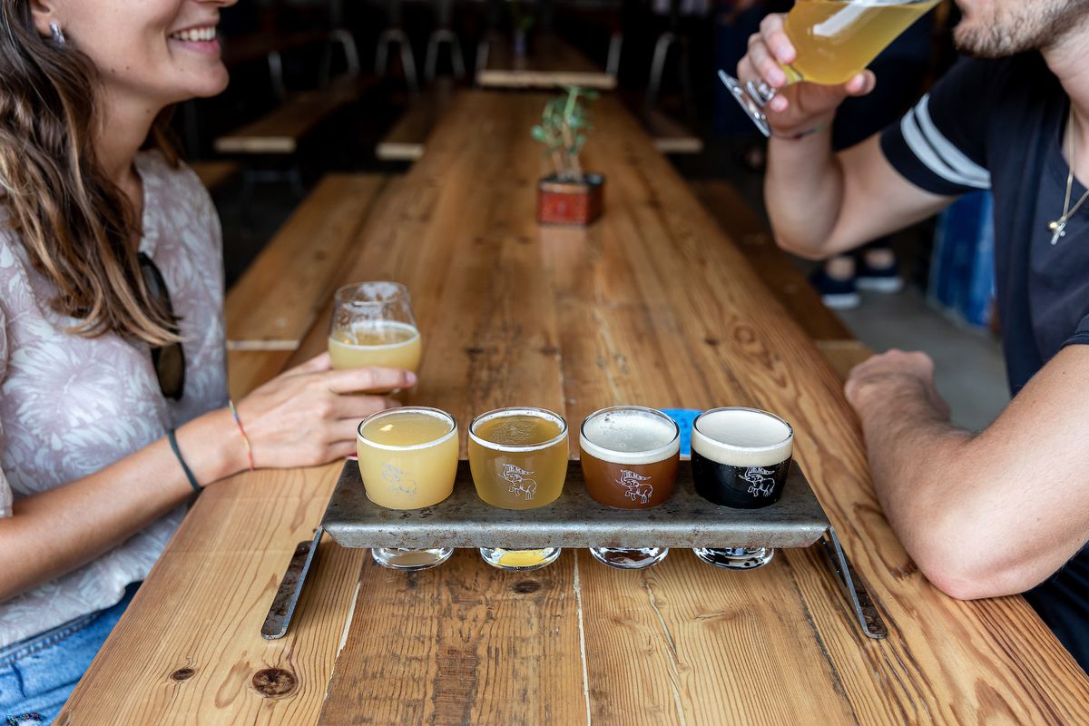 A series of beers in a metal tasting display shown from light to dark surrounded by two people sitting at a table.