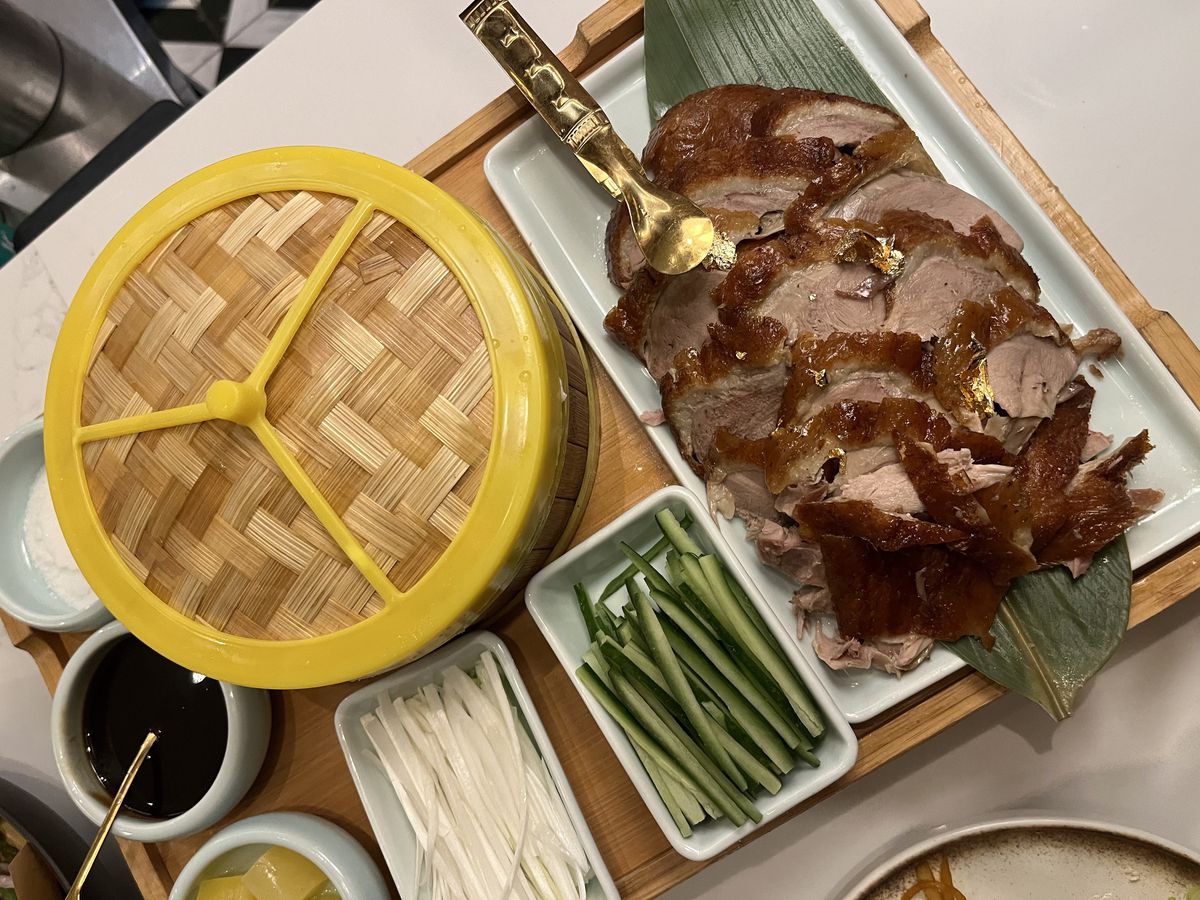 A platter of sliced roast duck with golden serving tongs plus side cups of julienned vegetables and dipping sauces, and a bamboo holder with thin pancake wrappers inside.