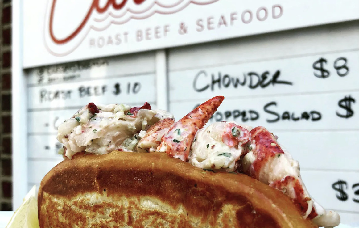 A hand holds a lobster roll in front of a sign with prices printed in black ink on white background. 