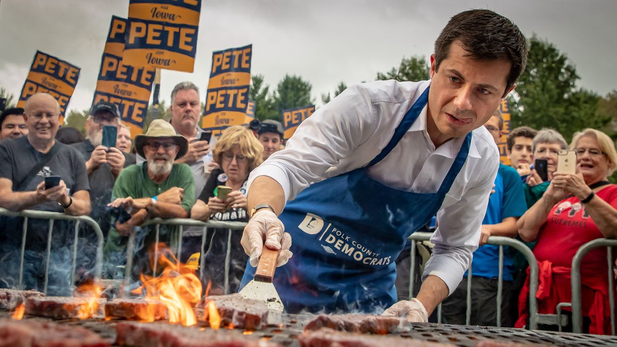 Pete Buttigieg grills a steak as supporters behind him hold signs and take photos.